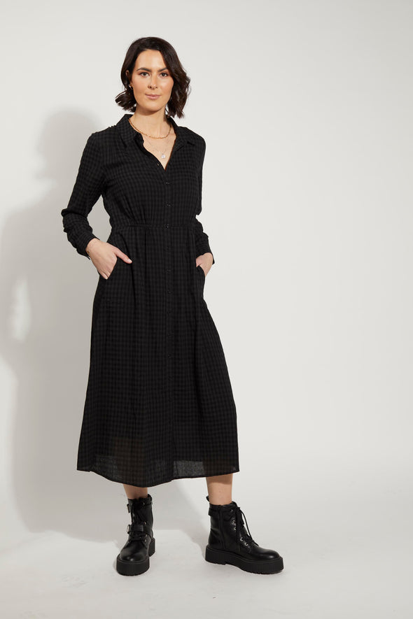The Canterbury Dress by Drama the Label. This stunning piece features a button-through design, complete with a sophisticated collar. The waist is elegantly adorned with elastic detailing, ensuring a flattering fit for all body types. With its midi length and long sleeves, complete with cuffs, this dress exudes timeless elegance. The classic Black Check pattern adds a touch of sophistication to any occasion.&nbsp;