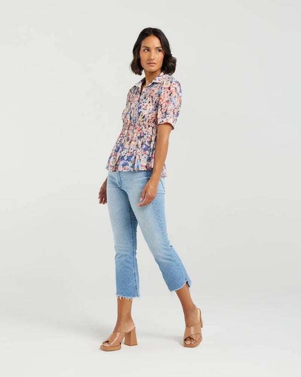 This short-sleeved shirt brings a playful touch with its softly puffed sleeves and delicate smocking that hugs the body. The gathered waist flares out into a subtle peplum, adding a flirty flair that flatters your silhouette. With refined tailoring and just the right amount of volume, it balances comfort and charm perfectly—ready to dress up your day or add fun to your casual look.